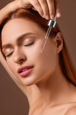 Güzel genç bir kadın yüzüne kozmetik serumu sürüyor, kahverengi arka planda, yakın plan