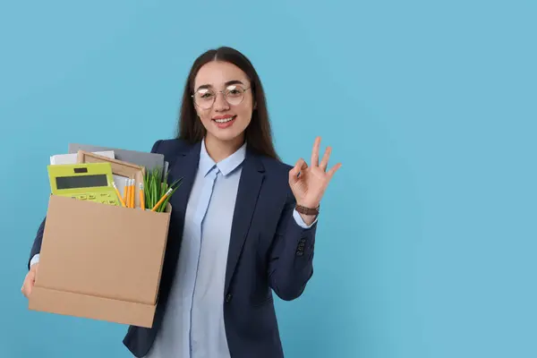 Happy unemployed woman with box of personal office belongings showing OK gesture on light blue background, space for text