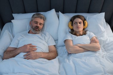 Evde yatakta horlayan kocasının yanında kulaklıkları olan sinirli bir kadın.