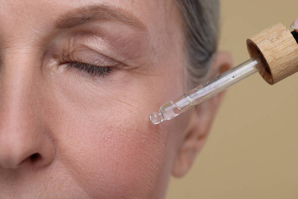 Senior woman applying cosmetic product on her aging skin against beige background, closeup. Rejuvenation treatment
