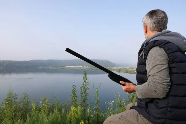 Man with hunting rifle sitting on wooden bench near lake outdoors. Space for text