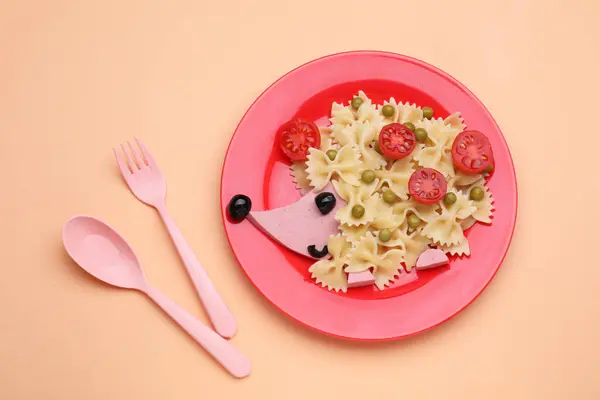 Plate with cute hedgehog made of delicious pasta, sausages and tomatoes on pale orange table, flat lay. Creative serving for kids