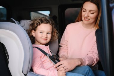 Anne kızını arabanın içindeki çocuk emniyet koltuğuna bağlıyor.