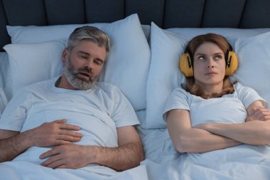 Evde yatakta horlayan kocasının yanında kulaklıkları olan sinirli bir kadın.