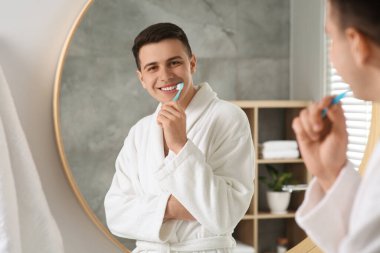 Adam banyoda aynanın yanında diş fırçasıyla dişlerini fırçalıyor.