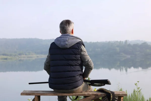 Man with hunting rifle sitting on wooden bench near lake outdoors, back view