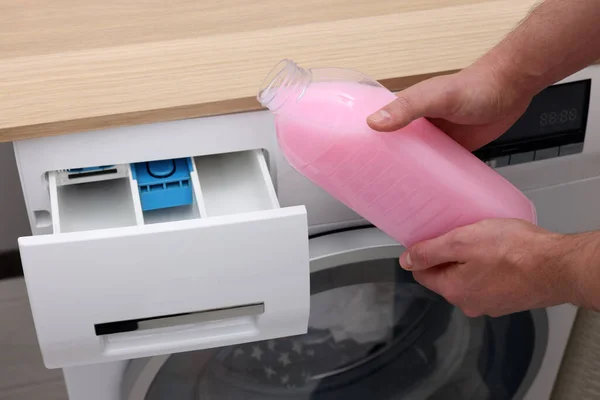 Man pouring fabric softener from bottle into washing machine indoors, closeup