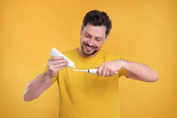 Happy man squeezing toothpaste from tube onto electric toothbrush on yellow background