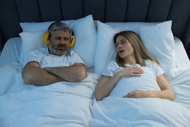 Evde yatakta horlayan karısının yanında kulaklıkları olan sinirli bir adam.