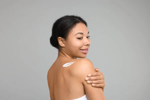 Young woman applying body cream onto back on grey background