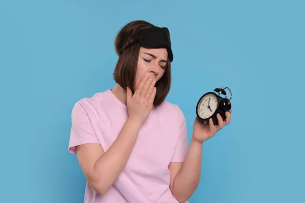 Sleepy young woman with sleep mask and alarm clock yawning on light blue background. Insomnia problem