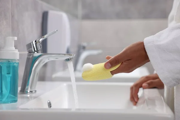 Young man cleaning face sponge above sink in bathroom, closeup