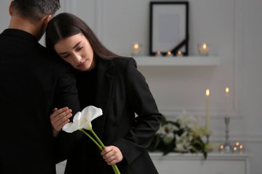 Sad couple with calla lily flowers mourning indoors, space for text. Funeral ceremony
