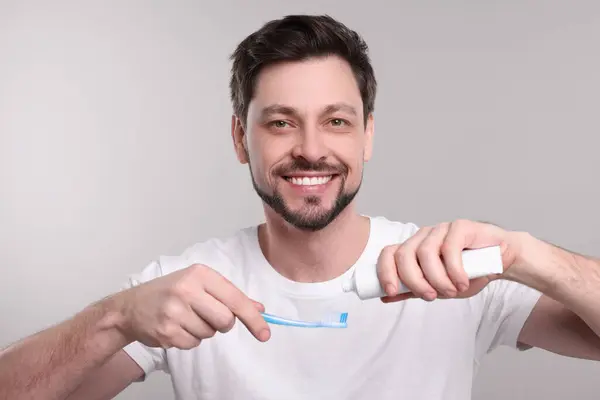 Happy man squeezing toothpaste from tube onto toothbrush on light grey background