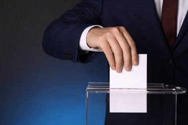 Man putting his vote into ballot box on dark blue background, closeup. Space for text clipart