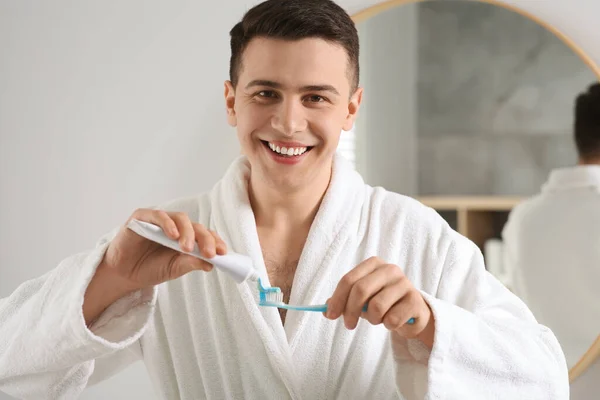 Happy man squeezing toothpaste from tube onto toothbrush in bathroom