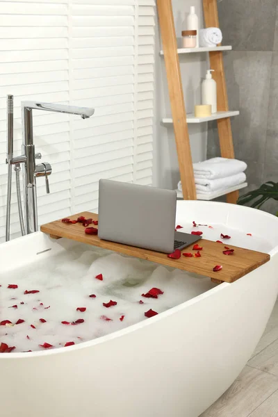 Wooden board with laptop and rose petals on bath tub in bathroom
