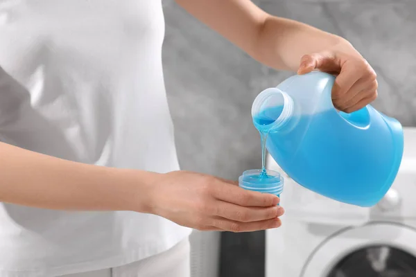 Woman pouring fabric softener from bottle into cap for washing clothes indoors, closeup