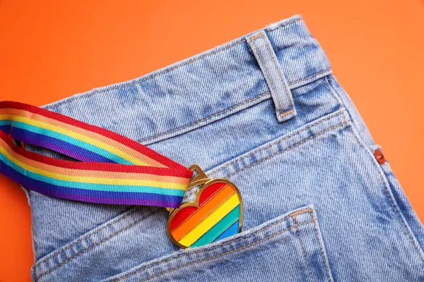 Jeans and rainbow ribbon with heart shaped pendant on orange background, top view. LGBT pride