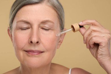 Yaşlanan cildine bej arka planda kozmetik ürünler uygulayan yaşlı bir kadın. Yenilenme tedavisi