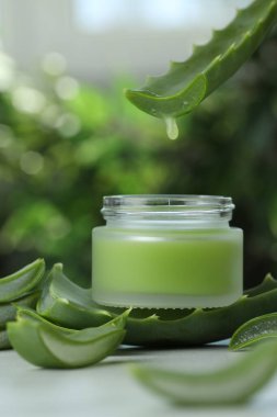 Aloe Vera suyu yapraktan kavanoza damlıyor. Beyaz masada bulanık arka planda krema var.