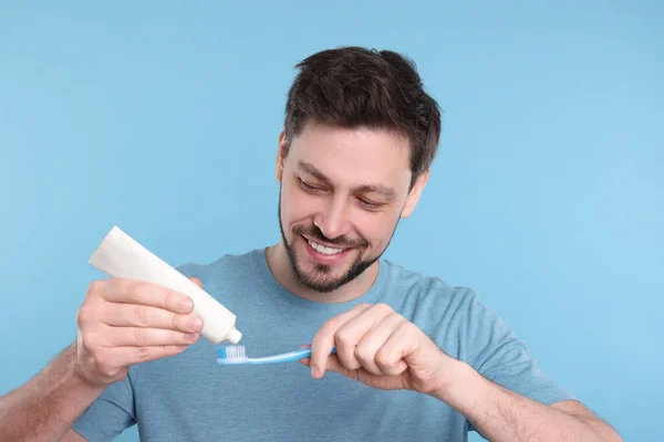Happy man squeezing toothpaste from tube onto plastic toothbrush on light blue background
