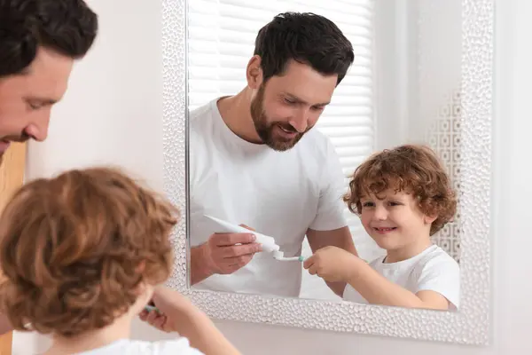 Father squeezing toothpaste from tube onto son`s toothbrush near mirror in bathroom