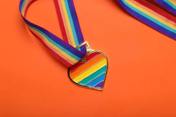 Rainbow ribbon with heart shaped pendant on orange background, top view. LGBT pride