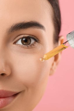 Pembe arka planda yüzüne serum süren güzel genç bir kadın. Yakın plan.