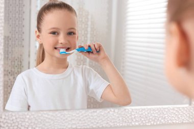 Küçük tatlı bir kız, içeride aynanın yanında dişlerini plastik diş fırçasıyla fırçalıyor.