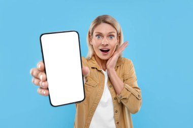 Açık mavi arkaplanda boş ekranlı akıllı telefonu tutan sürpriz bir kadın.