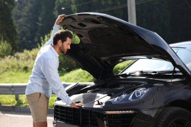 Stresli bir adam kırık bir arabanın kaputuna bakıyor.