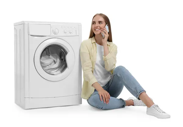 漂亮的年轻女子在洗衣机旁边用智能手机与白色背景的衣服交谈 — 图库照片