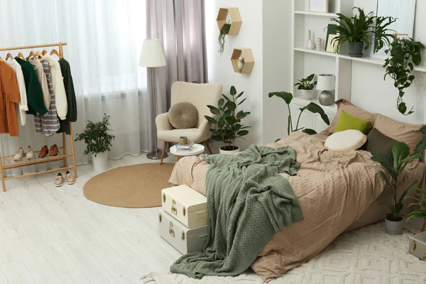 Stylish bedroom with comfortable bed, clothes rack and different houseplants. Interior design