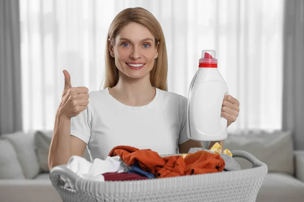 Woman holding fabric softener near basket with dirty clothes and showing thumbs up in room