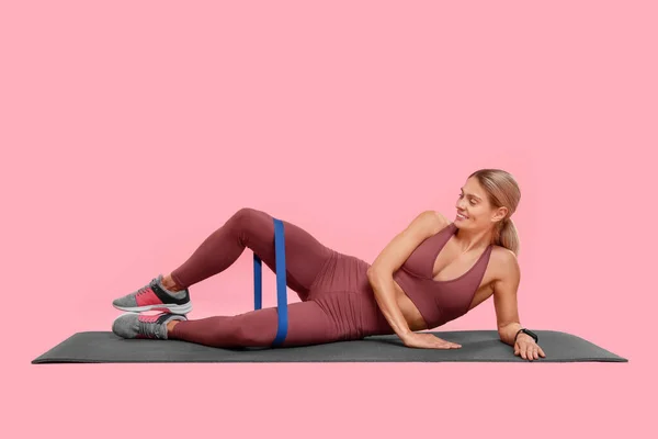 Woman exercising with elastic resistance band on fitness mat against pink background