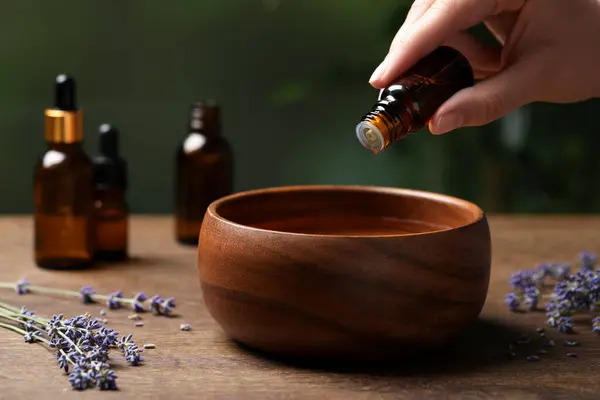 Woman dripping essential oil from bottle into bowl near lavender at wooden table, closeup. Space for text