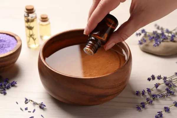 Woman dripping lavender essential oil from bottle into bowl at white wooden table, closeup