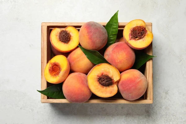Delicious juicy peaches and leaves in crate on light grey textured table, top view