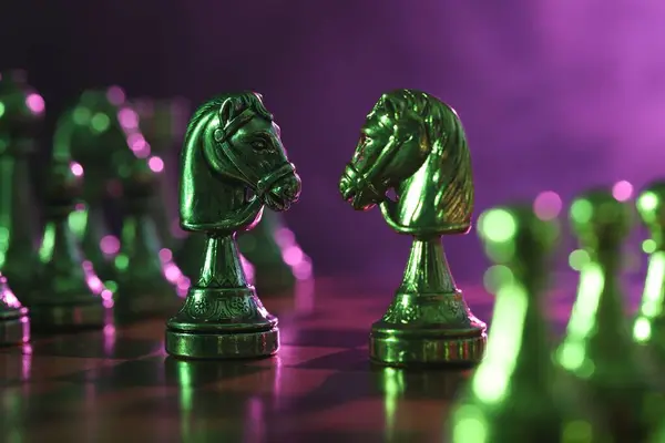 Two knights and other chess pieces on checkerboard in color light, closeup