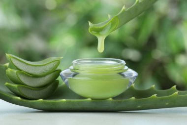 Aloe vera suyu yapraktan kavanoza damlıyor, beyaz masada krema ile yeşil bulanık arka planda, yakın plan