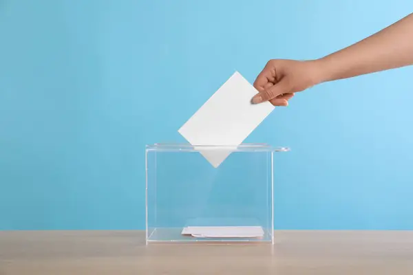 stock image Woman putting her vote into ballot box on wooden table against light blue background, closeup