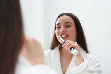 Banyoda aynanın yanında dişlerini elektrikli diş fırçasıyla fırçalayan genç bir kadın.