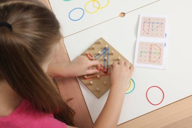 Motor skills development. Girl playing with geoboard and rubber bands at white table, above view clipart