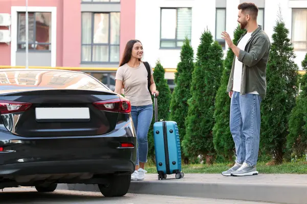 Long-distance relationship. Man waving to his girlfriend with luggage near car outdoors