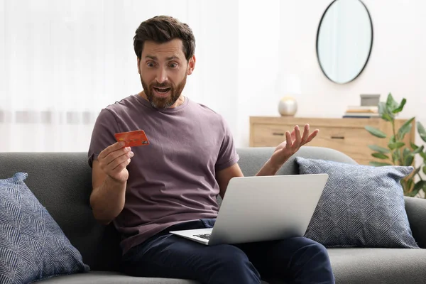Shocked man with credit card and laptop on sofa at home. Be careful - fraud