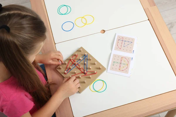 Motor skills development. Girl playing with geoboard and rubber bands at white table, above view