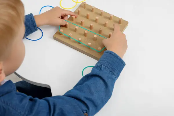 Motor skills development. Boy playing with geoboard and rubber bands at white table, closeup. Space for text