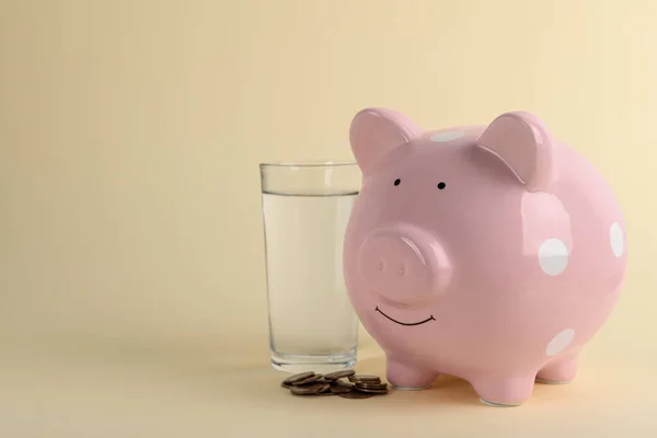 Water scarcity concept. Piggy bank, glass of drink and coins on beige background, space for text