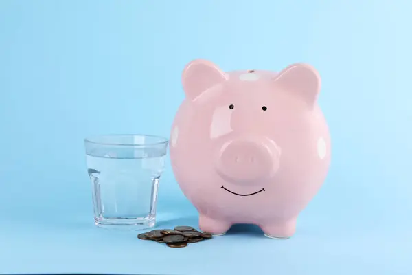 Water scarcity concept. Piggy bank, glass of drink and coins on light blue background